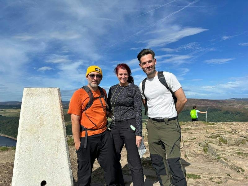 Three walkers smiling by a trig point on a sunny Peak District summit
