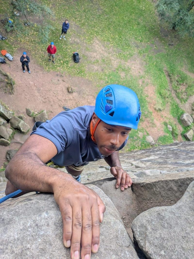 Climber in a blue helmet reaching the top of a gritstone route in the Peak District