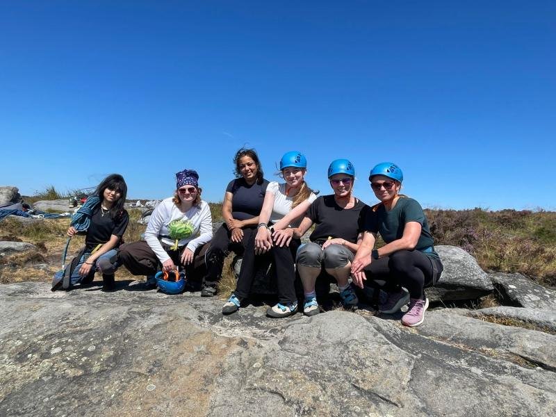 Group in climbing helmets sitting on gritstone boulders under a blue sky